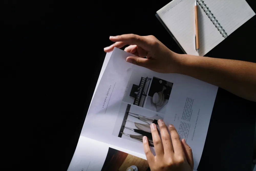 A person's hands holding an open magazine featuring design layouts, with a notepad and pen visible in the background on a dark surface.