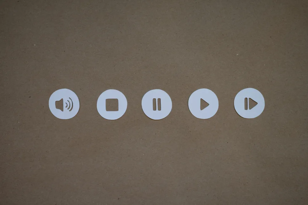 A row of five circular paper cutouts featuring media control icons: volume, pause, play, and two variations of play buttons, set against a brown background.