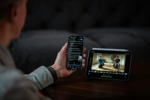 A person holds a smartphone while watching a video on a tablet. The setting features a cozy, modern interior.
